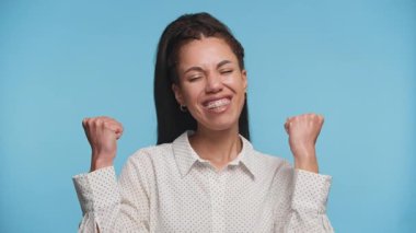 Neşeli, muzaffer bir pozda gülümseyen genç bir kadın yumruklarını kaldırmış, mavi arka planda başarı ve mutluluğu ifade ediyor..