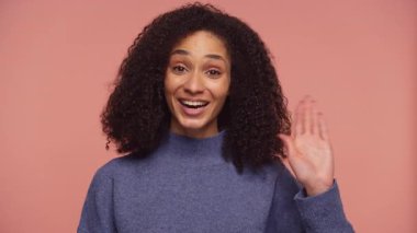 Gülümseyen ve elini sallayan mutlu genç bir kadın. Yumuşak pembe bir arka plan karşısında duruyor. Sevinç ve bağlılık ifade ediyor..