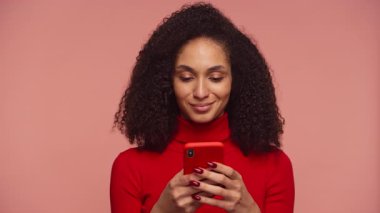 Kırmızılı genç kadın telefonuyla gülümsüyor, pembe arka planda dururken içeriğine mesaj atıyor veya göz gezdiriyor..