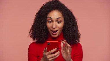 Akıllı telefonuna bakarken şaşkınlık ve heyecan gösteren genç bir kadın pembe arka planda neşe ve şaşkınlık ifade ediyor..