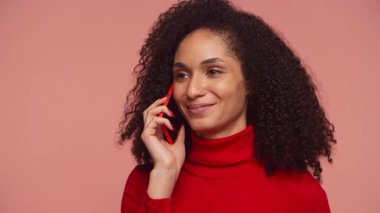 Bir telefon görüşmesi sırasında parlak bir şekilde gülümseyen neşeli genç bir kadın, kulağına pembe yumuşak bir arka plana karşı kırmızı bir akıllı telefon tutuyor..