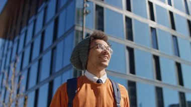 Sırt çantası ve günlük kıyafetleriyle mutlu genç adam güneşli bir günde cam gökdelende yürürken gülümsüyor..