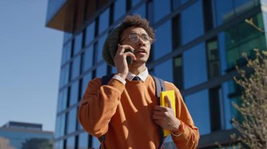 Sırt çantalı ve gündelik tarzda konuşan şehirli genç adam modern cam bir binanın önünde akıllı telefondan konuşuyor..