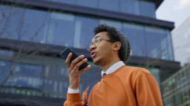 Gündelik kıyafetli genç bir öğrenci akıllı telefon tutuyor ve modern cam ofis binasının önünde dikilirken bir sesli mesaj kaydediyor..