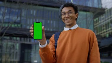 Elinde yeşil ekranlı akıllı bir telefon tutan mutlu genç öğrenci, şehirdeki modern bir ofis binasının önünde duruyor..