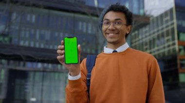 Açık havada gülümseyen bir öğrenci şehirdeki modern cam binaların yanında duran yeşil ekranlı bir akıllı telefonu hevesle işaret ediyor..