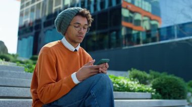 Akıllı telefonuyla merdivenlerde oturan düşünceli genç öğrenci şehirdeki modern cam ofis binasının yakınındaki bir mesaja cevap vermeyi düşünüyor..