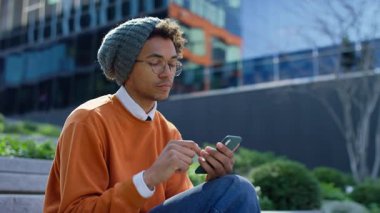 Şehir basamaklarında oturan genç adam, modern cam ofis binalarının yanındaki akıllı telefonuyla internete göz atmaya odaklanmış..