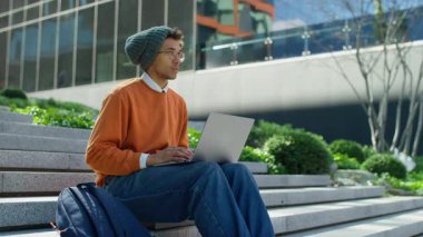 Modern ofis binalarının dışındaki basamaklara odaklanmış genç bir adam sıradan bir şehir ortamında dizüstü bilgisayarıyla çalışıyor..
