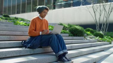 Modern binaların dışındaki basamaklarda oturan düşünceli öğrenci şehir ortamında dizüstü bilgisayarı üzerinde çalışırken fikirlerinden şüphe ediyor ve düşünüyor..