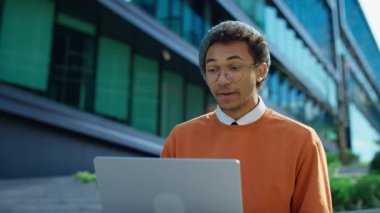Modern binaların önünde merdivenlerde oturan mutlu öğrenci güneşli bir şehirde dizüstü bilgisayarında neşeli bir video görüşmesi yapıyor..