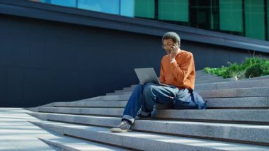 Şehir basamaklarında oturan sıradan bir genç adam, modern cam binaların yanında laptopunu kullanırken bir telefonla birden fazla iş yapıyor..
