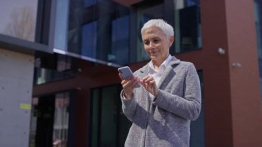 Modern binaların yakınında neşeli, son sınıf öğrencisi bir kadın. Akıllı telefonundan heyecan verici güzel haberler aldıktan sonra gülümsüyor ve kutluyor..