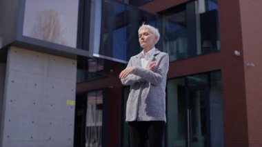 İş elbisesi giymiş kararlı yaşlı bir kadın modern bir ofis binasının önünde çapraz kollarla duruyor. Güç ve liderlik taşıyor..
