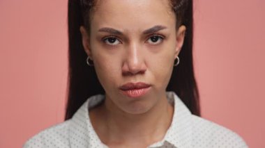 Kızgın, yoğun bir ifadeyle, pembe bir arka planda doğrudan kameraya bakan genç bir kadının yakın plan portresi..