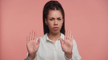 Ciddi bir ifade takınan genç bir kadın iki elini birden kaldırıyor ya da pembe bir arka plana karşı uyarı işareti yapıyor..