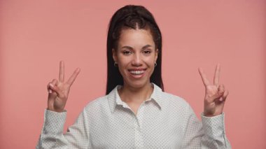 İki eliyle barış işaretleri gösteren gülümseyen genç bir kadın, yumuşak pembe bir arka plana karşı neşe ve pozitifliği ifade ediyor..