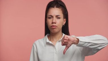 Ciddi bir genç kadın, pembe bir arka plana karşı hoşnutsuzluğunu ya da memnuniyetsizliğini ifade ederek, kaşlarını çatarak baş parmağıyla bir jest yapıyor..