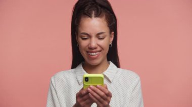 Akıllı telefonuyla mesajlaşırken gülümseyen mutlu genç bir kadın pembe stüdyo geçmişine karşı olumlu bir anın tadını çıkarıyor..