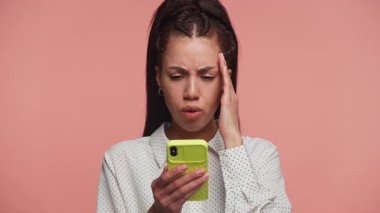 Endişeli genç bir kadın endişeli bir ifadeyle telefonuna bakıyor, kötü haberlere tepki veriyor veya pembe bir arka plana karşı rahatsız edici bir mesaj veriyor..