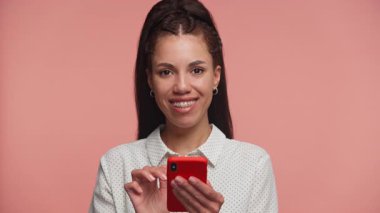 Mutlu genç bir kadın akıllı telefonunu parlak bir gülümsemeyle kullanıyor. Pembe stüdyo geçmişine karşı neşe ve heyecanla tepki veriyor..