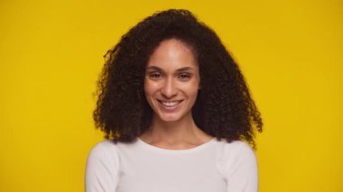 Kıvırcık saçlı neşeli genç bir kadın. Stüdyo ortamında parlak sarı bir arkaplan önünde poz veriyor..