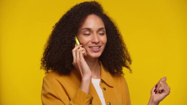 Kıvırcık saçlı neşeli genç bir kadın telefon görüşmesi yapıyor, canlı sarı bir arka planda dururken sıcak bir şekilde gülümsüyor..