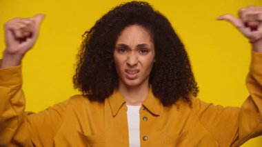 Kıvırcık saçlı etkilenmemiş genç bir kadın iki başparmağı aşağıda güçlü bir hoşnutsuzluk jesti yapıyor ve sarı bir arka plana karşı hoşnutsuzluğunu dile getiriyor..