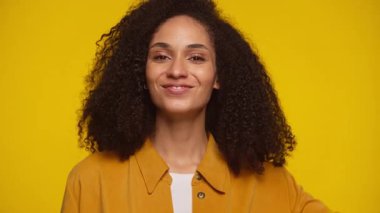 Kıvırcık saçlı mutlu genç bir kadın neşeli bir ifadeyle başparmak kaldırıyor, sarı bir arka plana karşı güvenle duruyor..