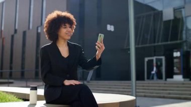 İnce çizgili takım elbiseli, kendine güveni tam bir kadın modern bir binanın önünde oturuyor. Akıllı telefonundaki bir video görüşmesi sırasında gülümsüyor..