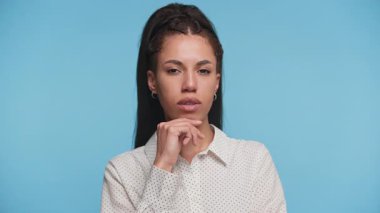 Özgüvenli bir kadının portresi düşünceli görünüyor, elleri çenesinde, beyaz bir gömlek giyiyor, düz mavi bir arka planı var..