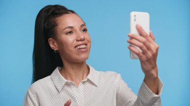 Telli ve noktalı tişörtlü genç bir kadın video görüşmesinde konuşuyor, akıllı telefonunu tutuyor ve kendini sıcak ve net bir şekilde ifade ediyor..