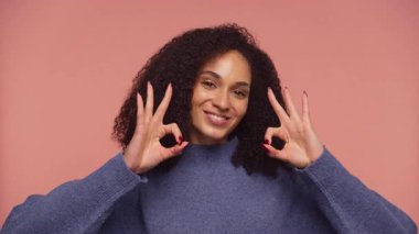 Gülümseyen ve her iki elini de gösteren neşeli bir kadın Tamam jest, pembe arka planda mavi bir süveter giyiyor.