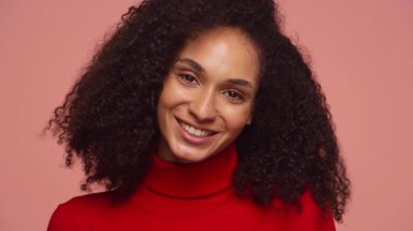 Kıvırcık saçlı, kameraya içten gülümseyen, yumuşak pembe arka planda kırmızı balıkçı yakalı bir kadın..