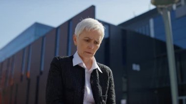 İş kıyafetleri içinde yaşlı bir kadın dışarıda oturuyor. Ofis binalarının yanında dizüstü bilgisayar kullanırken yüzünü strese sokuyor..