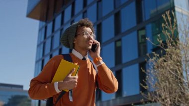 Gözlüklü ve sırt çantalı genç bir adam, elinde defterler var ve modern bir şehir binasının önünde telefonla konuşuyor, muhtemelen bir öğrenci..