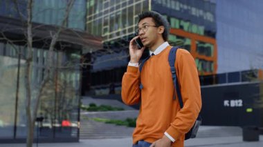 Sırt çantalı ve bereli kendinden emin bir erkek öğrenci modern şehir kampüsünde yürürken akıllı telefondan konuşuyor..