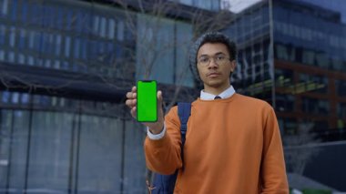 Elinde yeşil ekran görüntüsü olan turuncu kazaklı genç bir öğrenci, modern binanın önünde duruyor, uygulama modeli veya tasarımı için hazır..