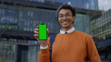 Portakal rengi süveter giyen neşeli öğrenci yeşil ekranlı akıllı telefonu modern cam binanın önünde sergiliyor. Uygulama modeli için mükemmel..