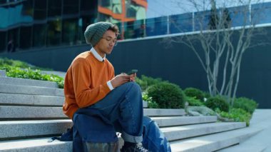 Modern bir üniversite ortamında rahat ve odaklanmış bir şekilde dışarıda oturmuş akıllı telefondan mesaj atıyor..