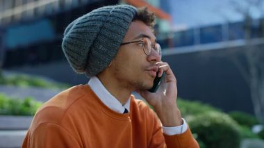 Gözlüklü ve bereli genç bir adamın modern bir binanın önünde merdivenlerde otururken telefonla konuşurken yan görüntüsü..