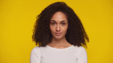 Kendine güvenen kıvırcık saçlı, cesur sarı arka planda sakince kameraya bakan genç bir kadının stüdyo portresi..