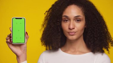 Genç bir kadın güvenli bir şekilde yeşil ekran ekranlı akıllı bir telefonu sunuyor. Parlak sarı bir arkaplan karşısında model ya da uygulama gösterileri için mükemmel..