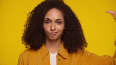 Sarı arka planda şüpheli bir ifadeyle baş parmak işareti yapan kıvırcık saçlı kadın portresi, memnuniyetsizlik veya hoşnutsuzluğu ifade ediyor..