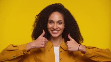 Kıvırcık saçlı, gülümseyen kadın sarı arka planda iki baş parmağını gösteriyor. Pozitif, onaylı ve neşeli enerjiyle cesaret veriyor..