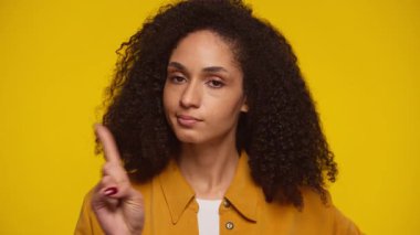 Sarı tişörtlü kıvırcık saçlı kadın işaret parmağıyla uyarı hareketi yapıyor. Sarı arka planda dikkat ya da hoşnutsuzluk gösteriyor..