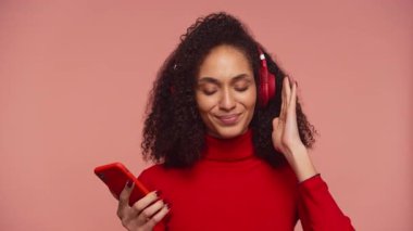 Kıvırcık saçlı, kırmızı kazaklı, gözleri kapalı müzik dinleyen, akıllı telefon tutan ve pembe arka planda kırmızı kulaklık takan bir kadın..