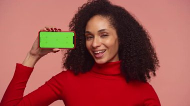 Kırmızı balıkçı yakalı mutlu kıvırcık saçlı kadın yeşil ekranlı akıllı telefonu tutuyor ve pembe arka planda poz veriyor..