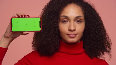 Kırmızı kazaklı kıvırcık saçlı kadın elinde yeşil ekranlı akıllı telefon ve onaylamadığı için başını sallayarak pembe arka plana karşı memnuniyetsizliğini dile getiriyor..