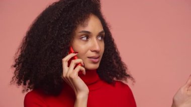 Kıvırcık saçlı, kırmızı balıkçı yakalı kadın telefon görüşmesi yapıyor, akıllı telefonu kulağına dayayıp, ilgiyle yan tarafa bakıyor..